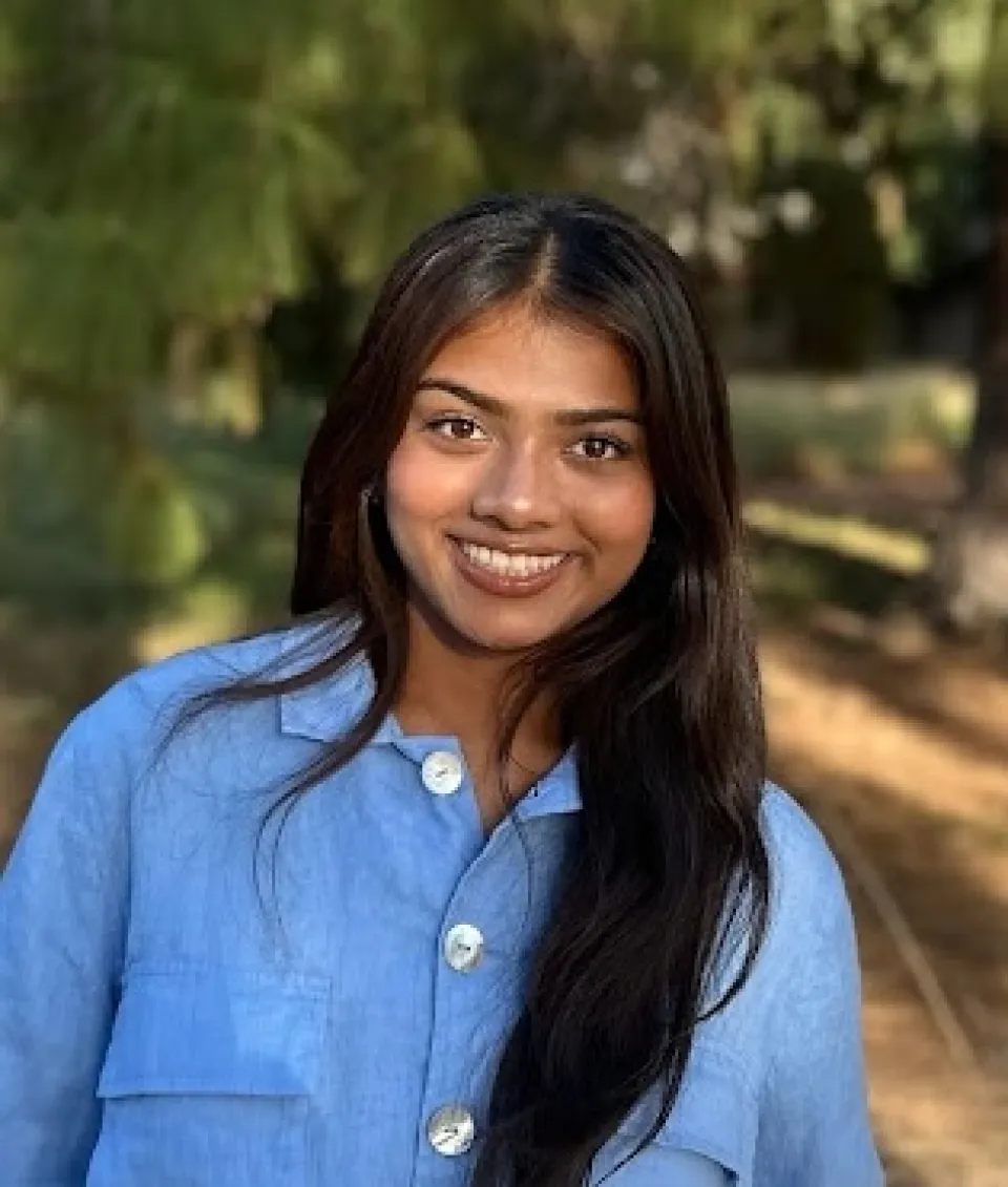 A woman with long dark hair, wearing a light blue button-up shirt, smiles outdoors with trees and sunlight in the background.
