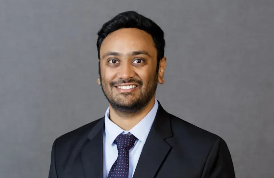 Man wearing a black suit, white shirt, and patterned tie stands in front of a plain gray background, smiling at the camera.