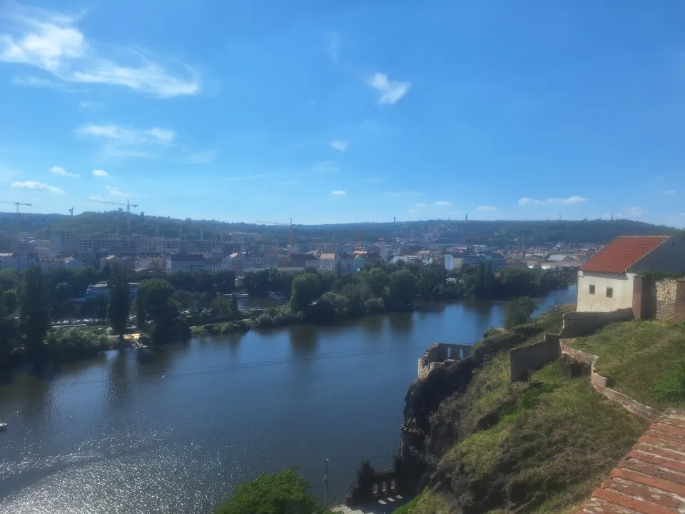 A river runs through a city with buildings and cranes, bordered by greenery and a hillside with a small structure under a blue sky.
