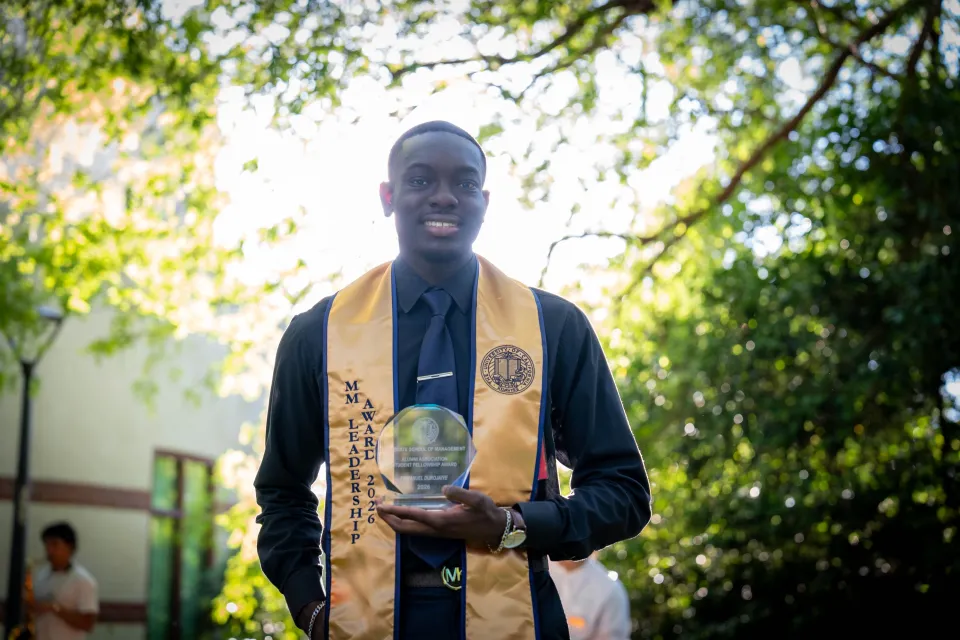 A person in a suit wears a gold sash and holds an award plaque while standing outdoors with sunlight filtering through trees.