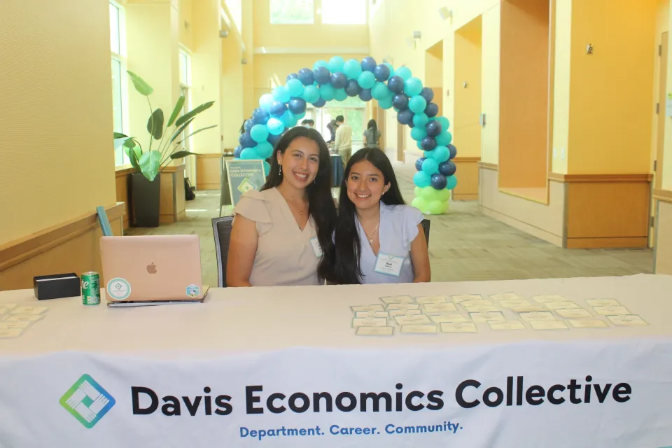 Two women sit behind a table with “Davis Economics Collective” banner in a bright hallway, with name tags, a laptop, and a blue-green balloon arch in the background.