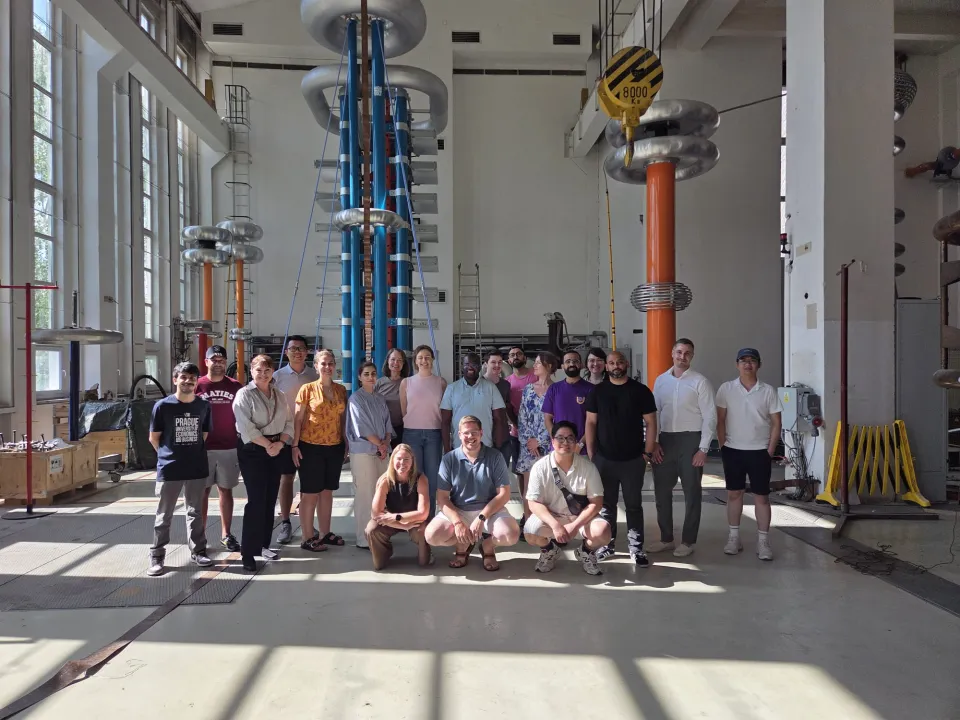 A group of people pose for a photo inside a large industrial laboratory with tall electrical equipment and high ceilings.