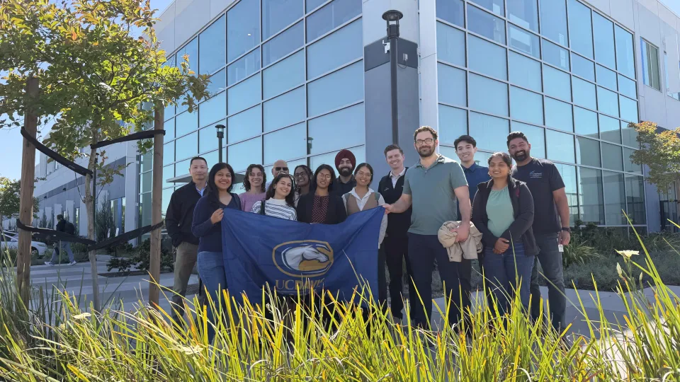 A group of people stands outdoors in front of a modern building, holding a blue flag with the UC Davis logo.