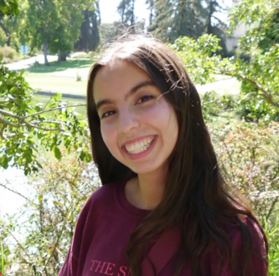 Renata Rosales-Verdugo smiles outdoors. She is wearing a maroon shirt and is standing in front of green trees and sunlight.