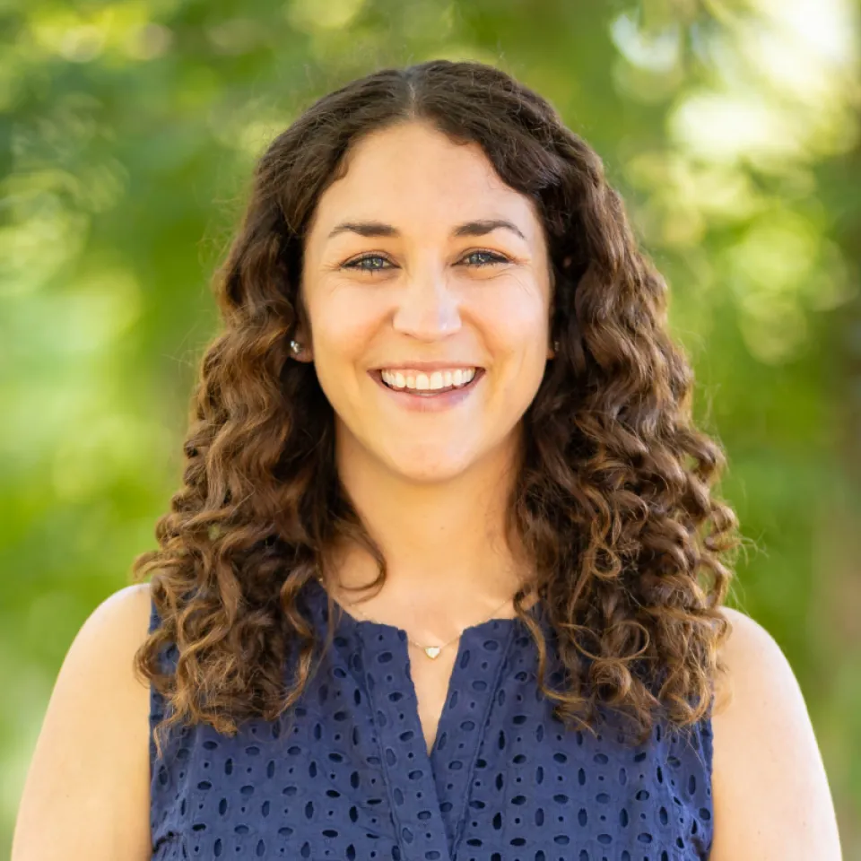 Woman with curly brown hair wearing a sleeveless navy blue top smiles outdoors in front of a blurred green background.