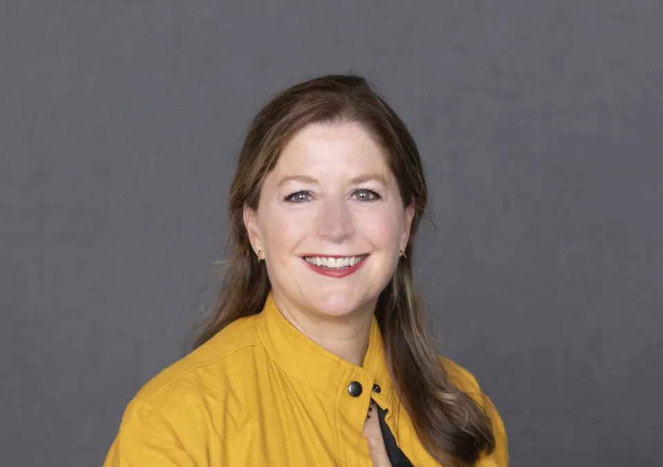 Anne Retterer wearing a mustard yellow jacket, smiles at the camera against a plain gray background.
