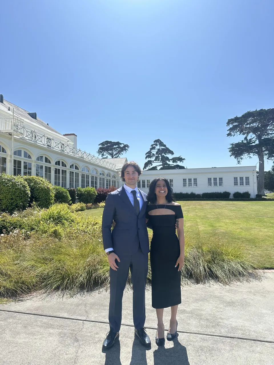 Two people in formal attire stand smiling outside a large white building on a sunny day, with greenery and trees in the background.
