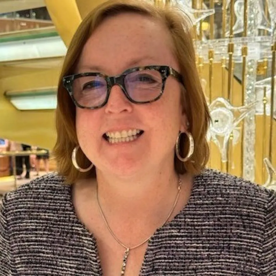 Woman with short red hair, glasses, and hoop earrings smiles indoors. She wears a gray textured top and a silver necklace, with a golden and glass background behind her.