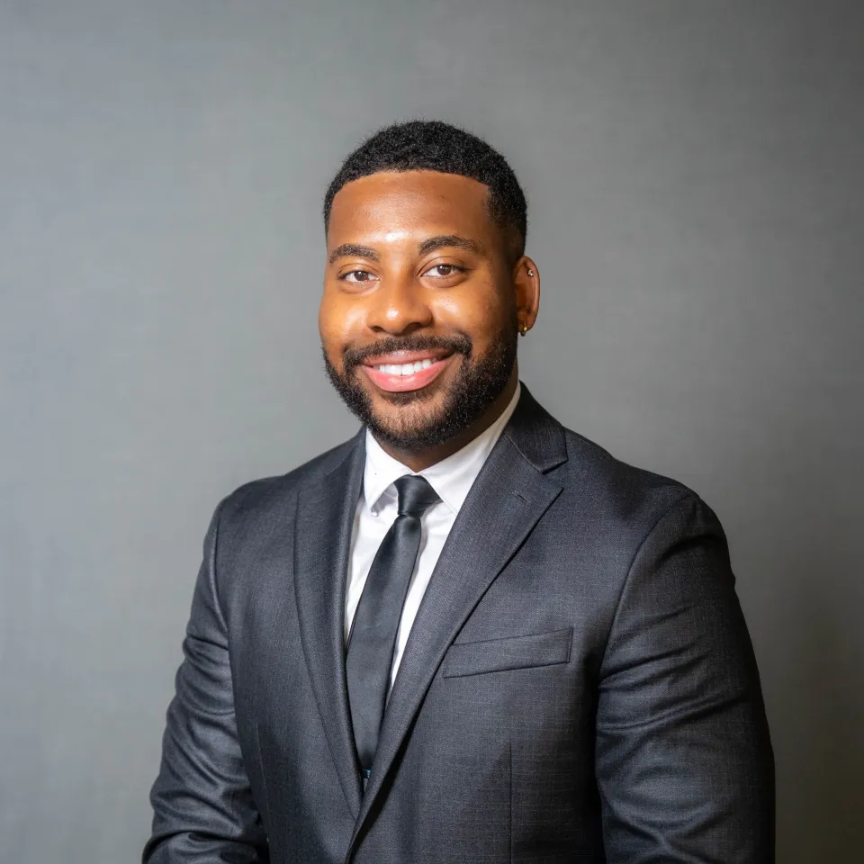 Man in a dark suit, white shirt, and black tie smiling while posing in front of a plain grey background.