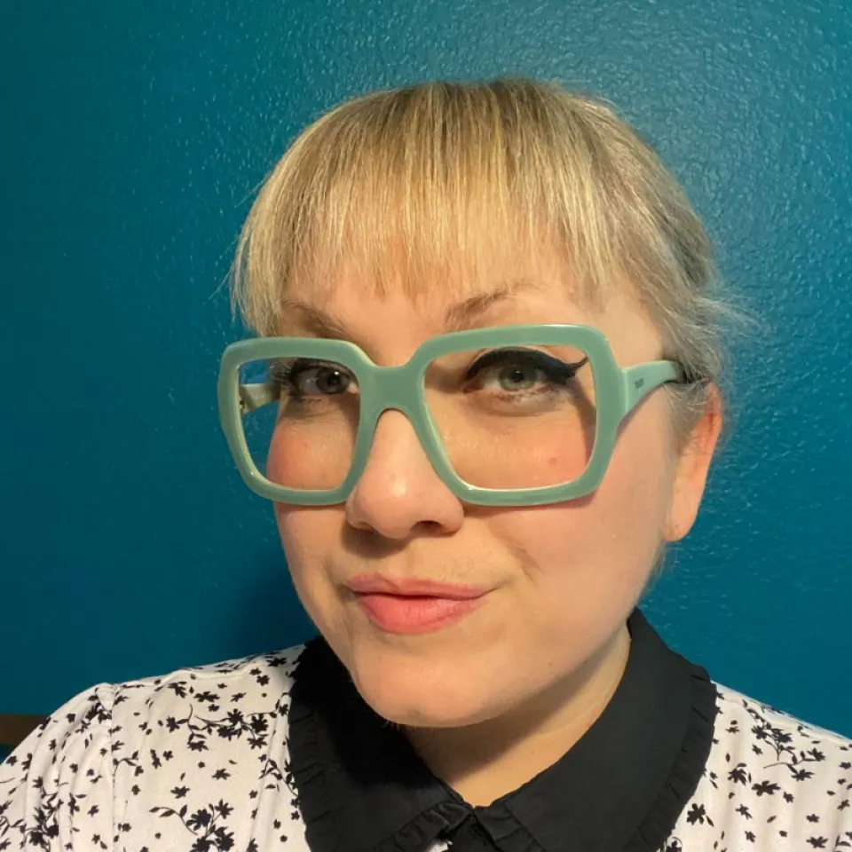 Person with blonde hair, large blue glasses, and winged eyeliner poses in front of a teal wall, wearing a white shirt with black patterns and a black collar.