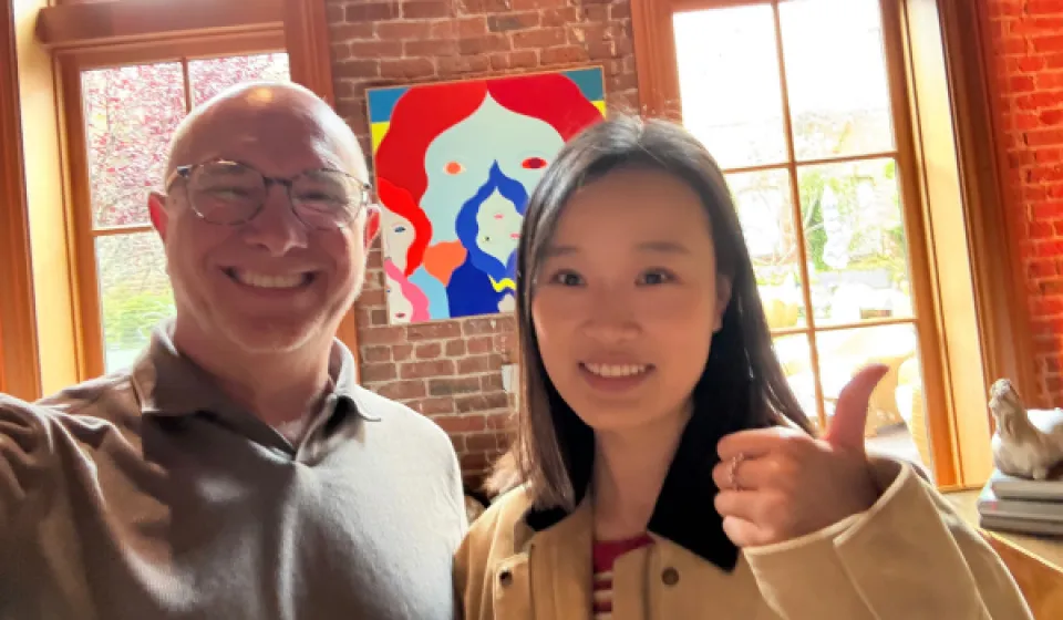 Two people smile for a photo indoors, with a colorful painting and large windows in the background. One person gives a thumbs-up gesture.