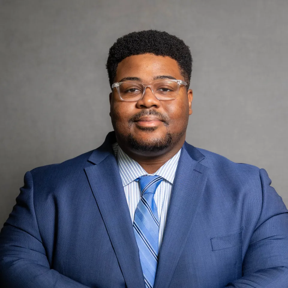 A man wearing glasses, a blue suit, striped shirt, and blue tie stands in front of a plain gray background, facing the camera with a neutral expression.