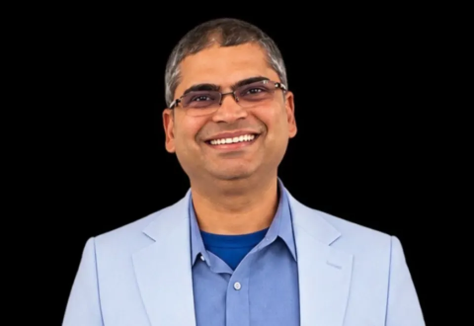 A man wearing glasses, a light blue blazer, and a blue shirt is smiling in front of a plain black background.