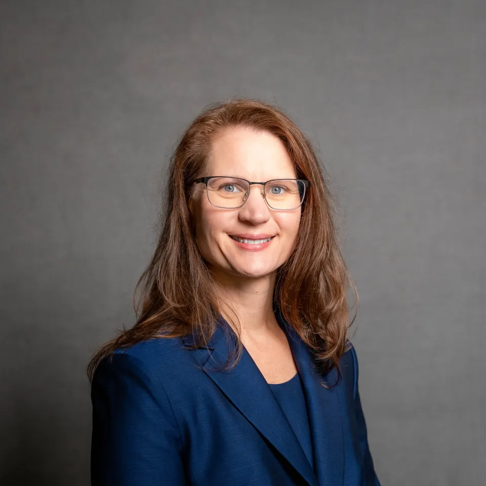 A woman with long brown hair and glasses wearing a blue blazer smiles at the camera against a plain gray background.