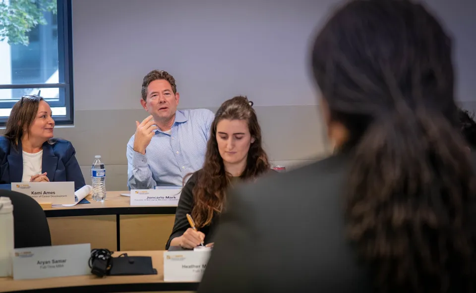 A man speaks during a classroom discussion while two women listen and take notes. Nameplates and notebooks are on the desks.