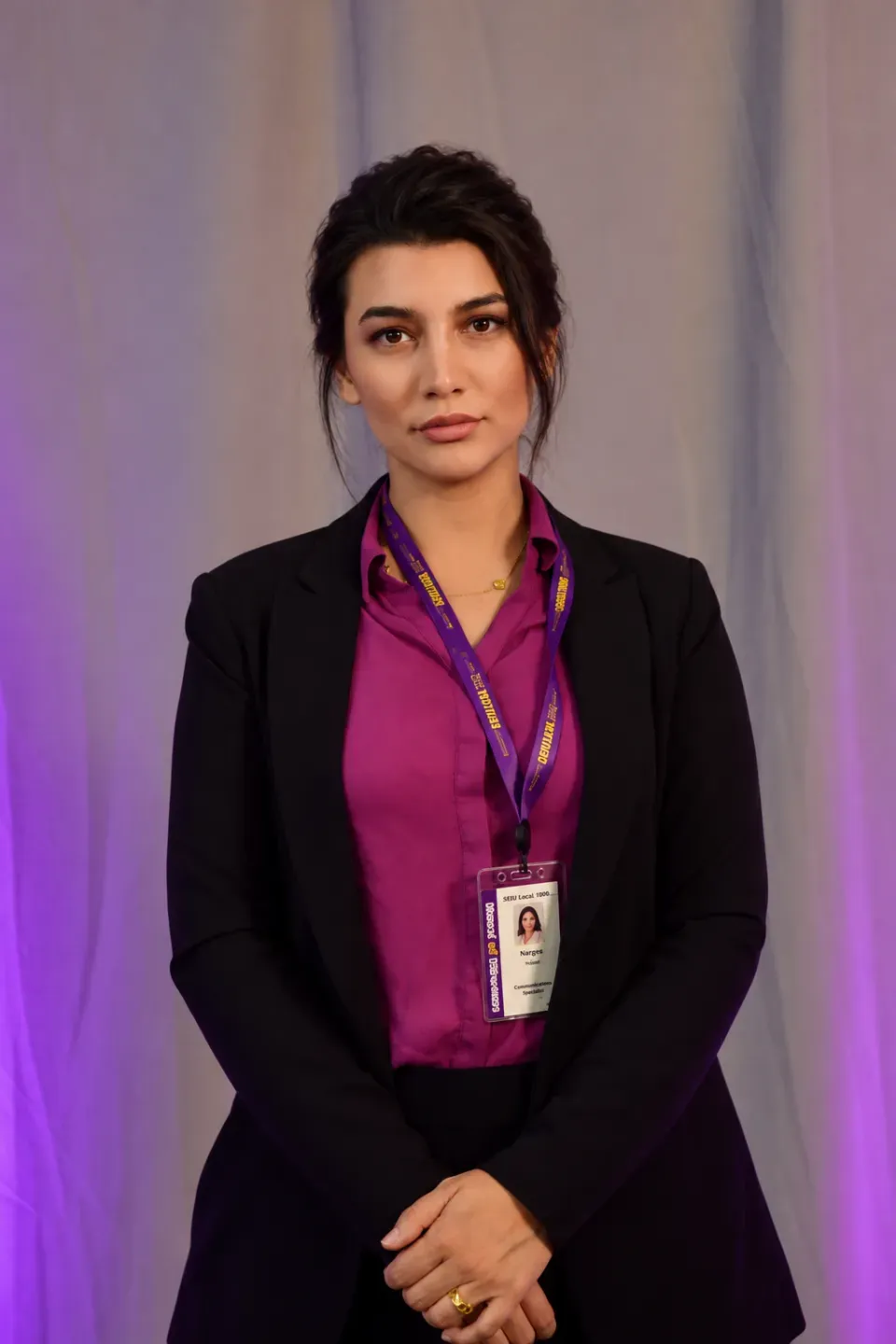 A woman in a black blazer and purple shirt stands in front of a gray curtain, wearing a lanyard with an ID badge around her neck.