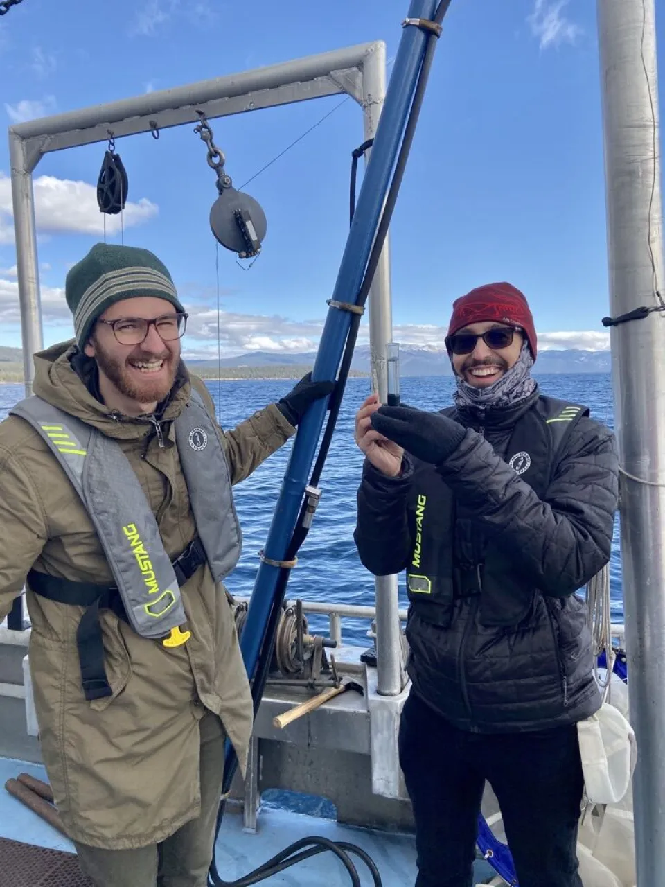 Two people in cold-weather gear and life jackets stand on a boat holding scientific equipment, with water and sky in the background.