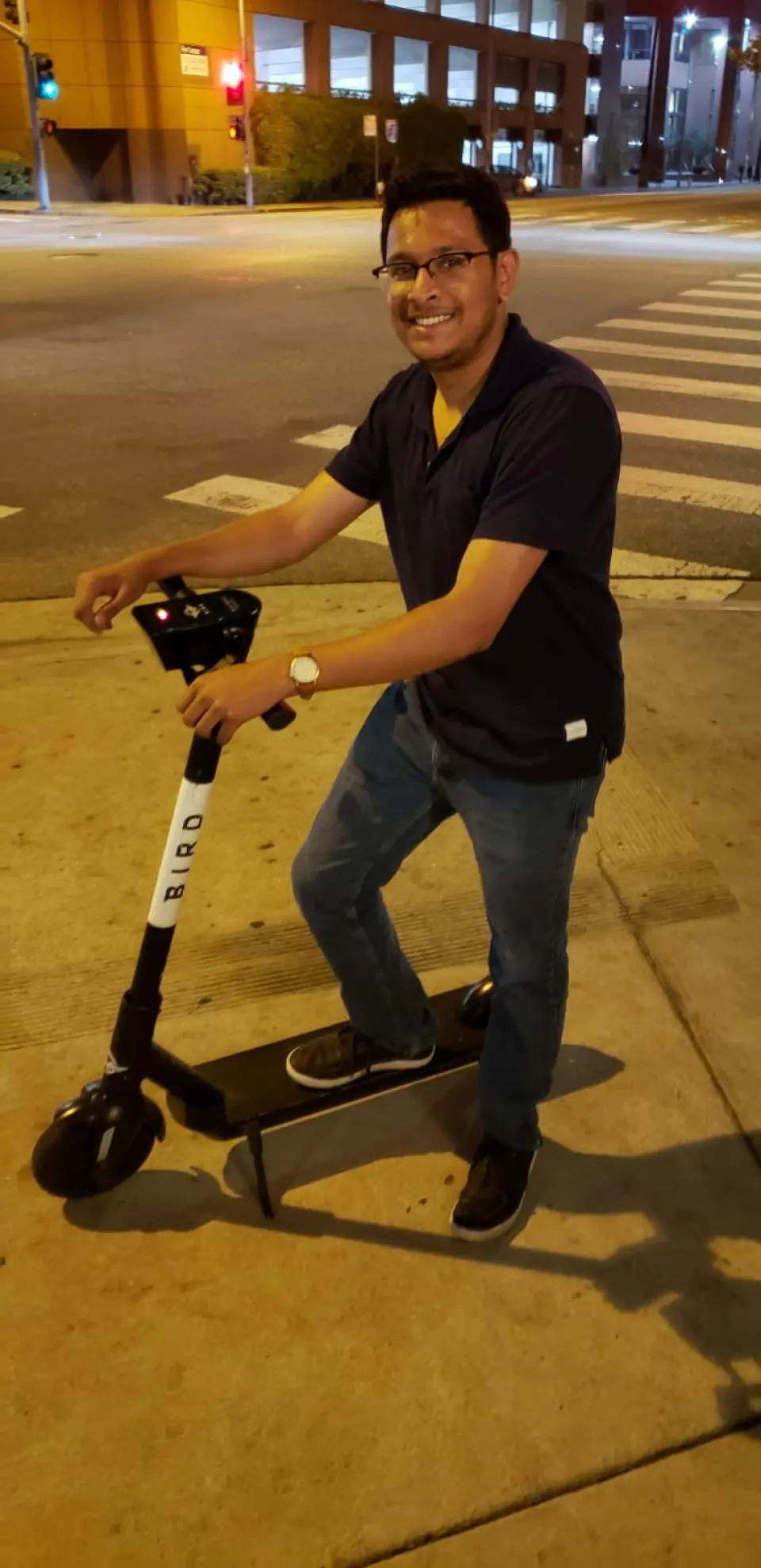 A man wearing glasses, a navy shirt, and jeans smiles while standing with one foot on a Bird electric scooter on a city sidewalk at night.