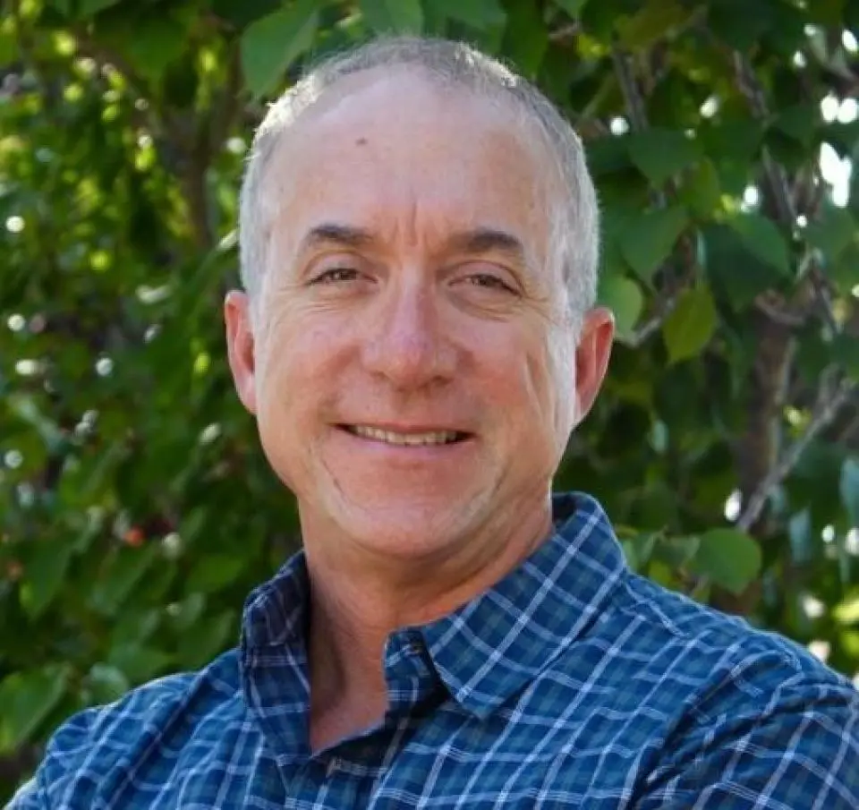 A middle-aged man with short gray hair smiles while standing outdoors in front of green leafy trees, wearing a blue plaid shirt.