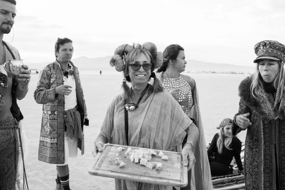 A woman wearing large horn-shaped accessories and sunglasses holds a wooden tray with food, surrounded by people in eclectic clothing on a desert landscape.