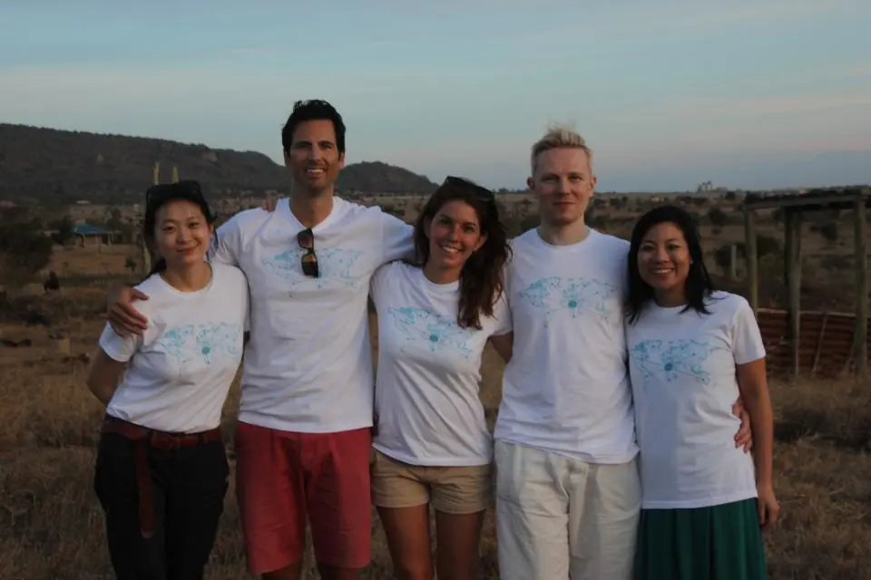 Five people stand together outdoors in a dry, grassy landscape, all wearing matching white t-shirts with a blue graphic design.
