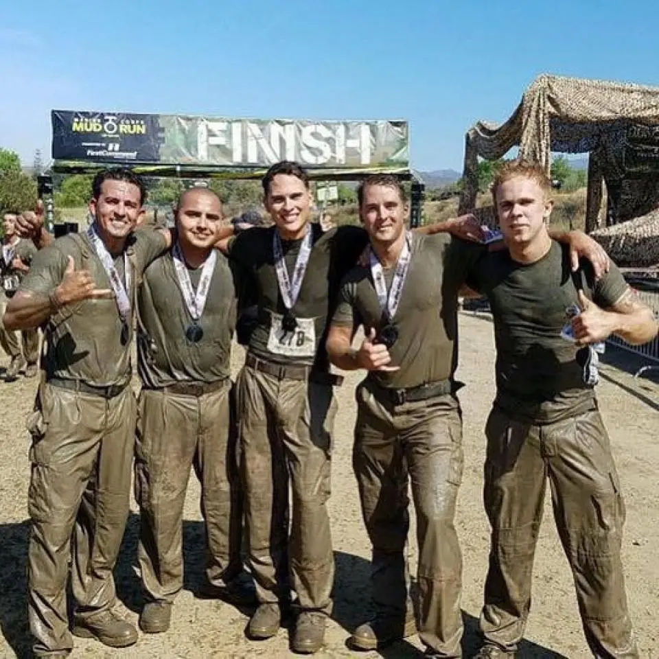 Five men stand together, covered in mud, smiling and posing in front of a finish line banner after completing an outdoor mud run race.