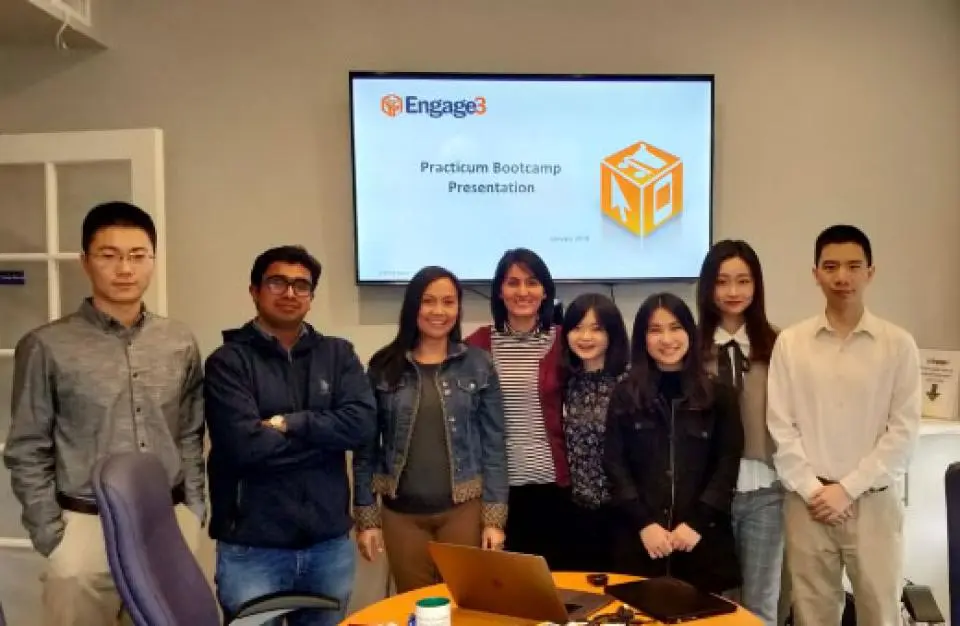 Eight people stand in front of a screen displaying "Engage3 Practicum Bootcamp Presentation" in an office setting.