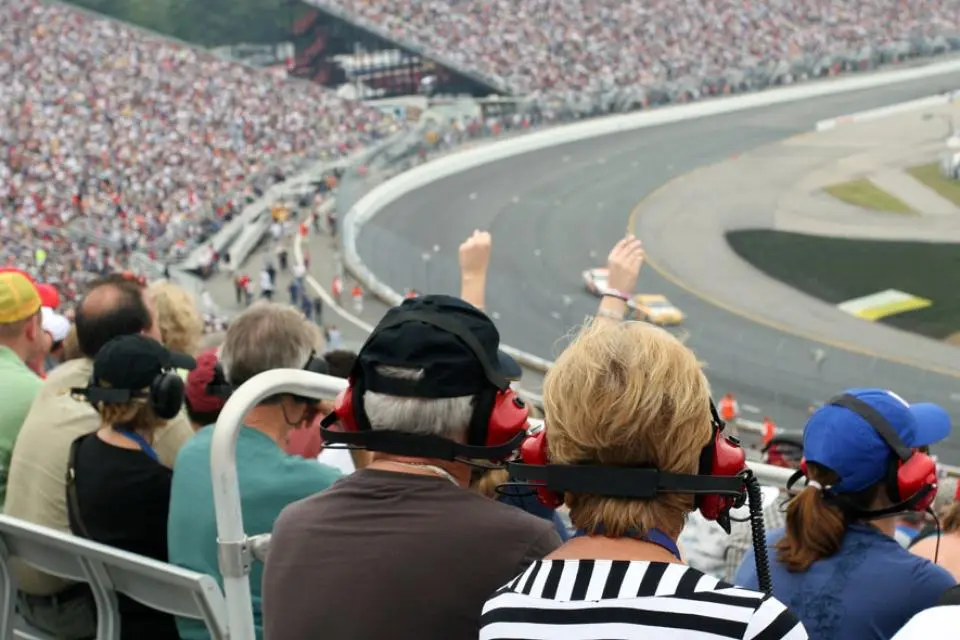 Spectators wearing headphones watch a car race at a crowded stadium, with cars speeding around a curved track in the background.
