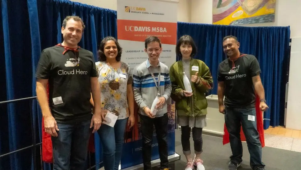 Five people stand in front of a UC Davis MSBA banner, two wearing "Cloud Hero" shirts and capes, posing for a group photo indoors.