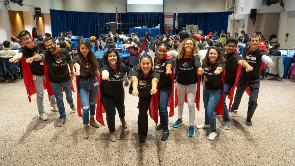 A group of people wearing matching Cloud Hero shirts and red capes pose with arms outstretched in front of a seated audience in a large event space.