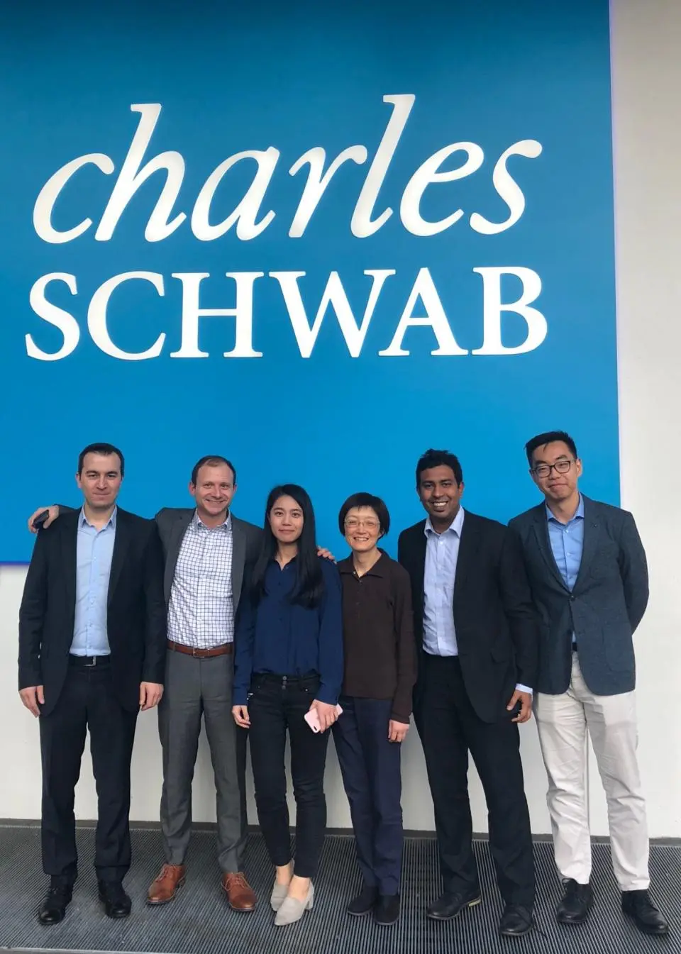 Six people stand in front of a large Charles Schwab sign, posing for a group photo.