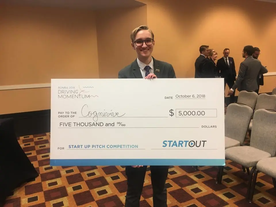A person in a suit holds a large ceremonial check for $5,000 made out to "Cognivive" for winning the StartOut pitch competition, dated October 6, 2018.