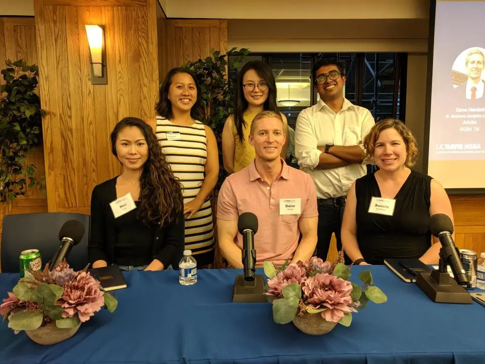 Six people pose for a group photo at a conference panel table, with name tags and microphones in front of them; floral arrangements and drinks are on the table.