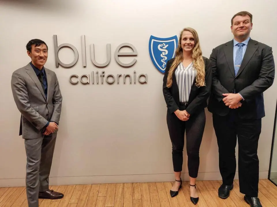 Three people in business attire stand in front of a wall with the Blue Shield of California logo and text.