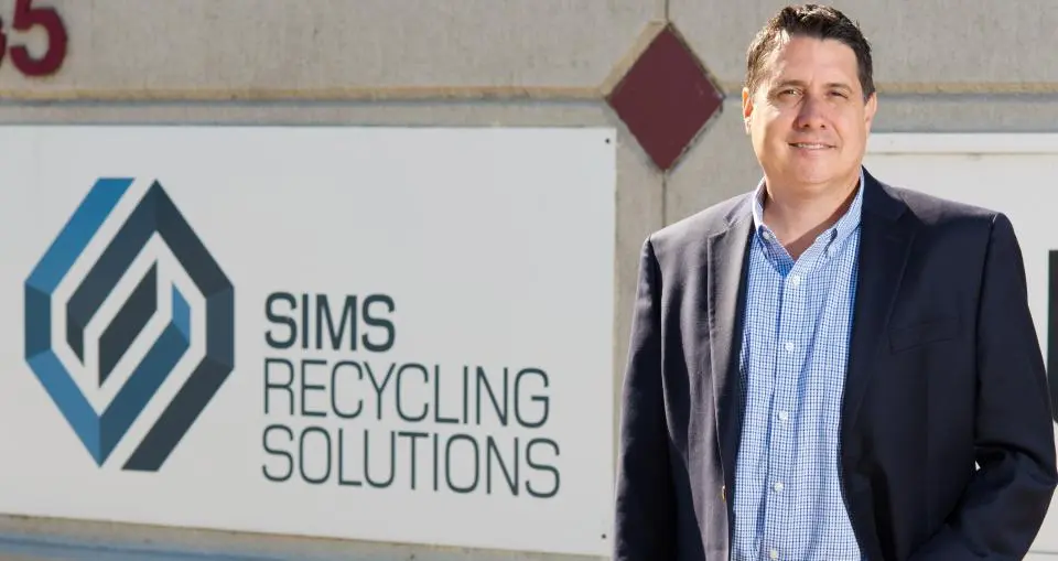 A man in a suit stands in front of a wall sign that reads "Sims Recycling Solutions" with the company logo.