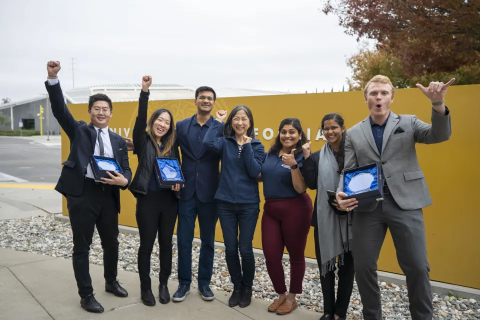 Group of individuals standing in front of the UC Davis sign with their hands raised celebrating