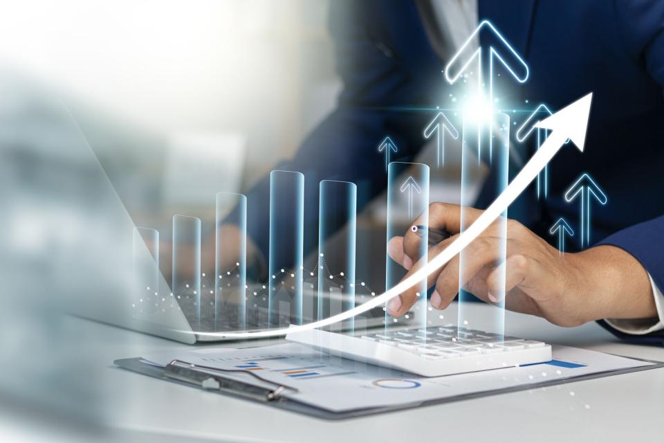 Business professional analyzing financial data on a laptop with a calculator, accompanied by a digital overlay of upward-trending bar graphs and arrows representing growth and success.