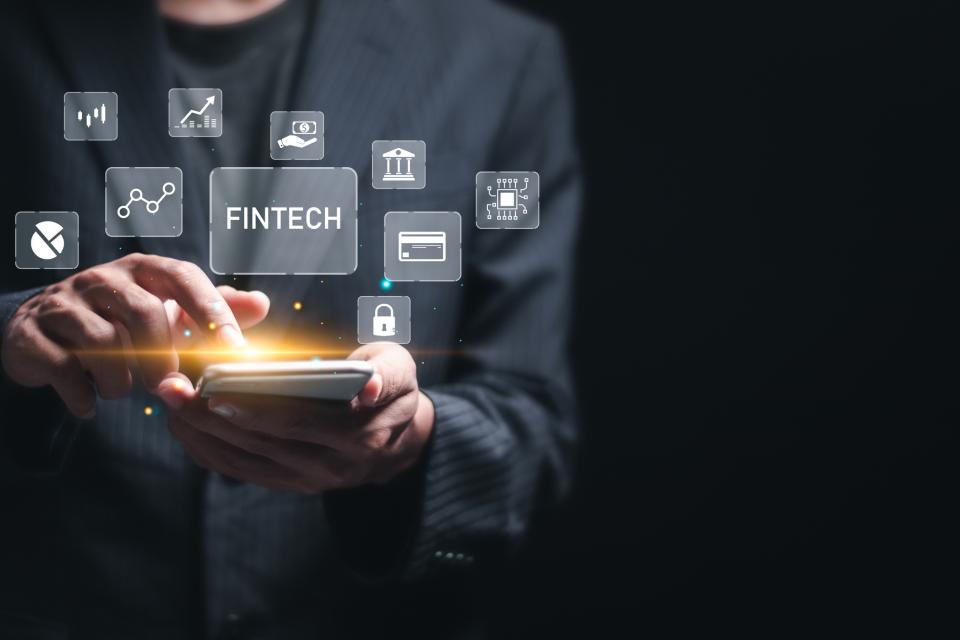 Person in a business suit using a smartphone, with floating digital icons representing financial technology (fintech) services such as data charts, banking, digital payments, and cybersecurity.