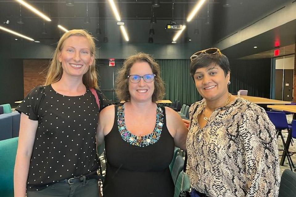 Jaimie Asakawa and two women stand together smiling inside a conference-style room with colorful chairs and tables in the background.
