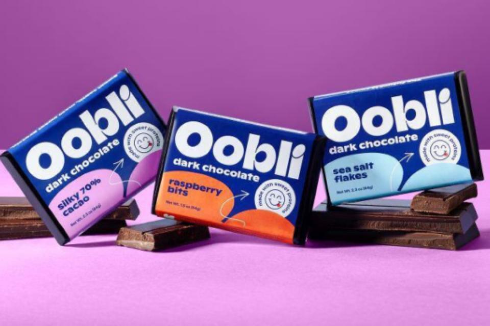Oobli dark chocolate bars in Silky 70% Cacao, Raspberry Bits, and Sea Salt Flakes flavors displayed on chocolate squares against a purple background.