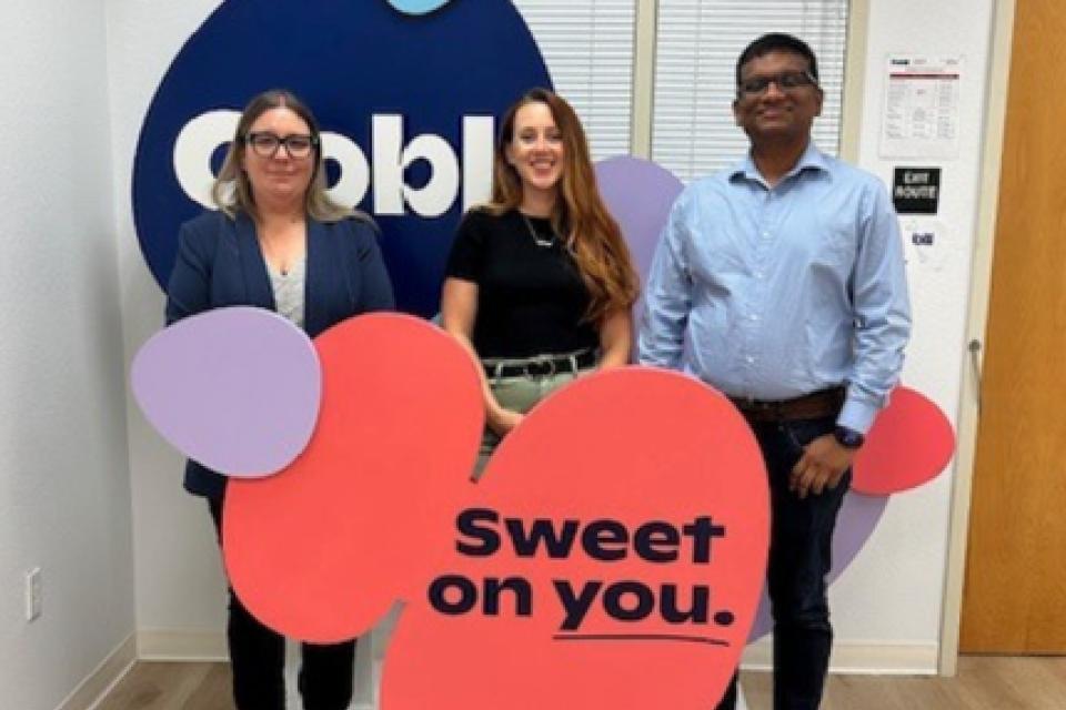Three people stand in front of an Oobli display sign inside an office, holding colorful cutouts with the phrase “Sweet on you.”