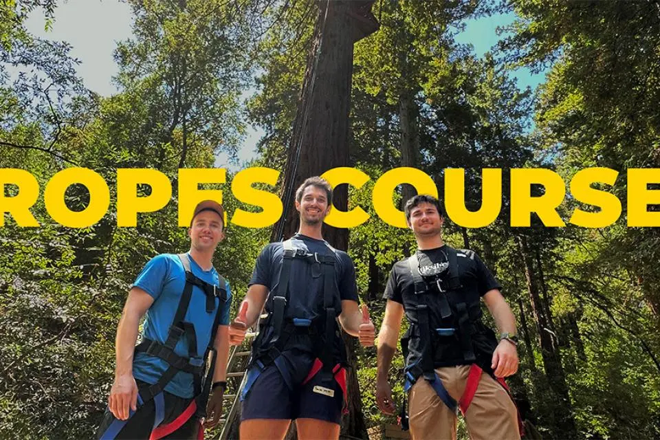 Three UC Davis MBA students wearing harnesses pose together at the Challenge Sonoma Ropes Course in Glen Ellen, California, standing in front of tall redwood trees with the words “ROPES COURSE” in bold yellow behind them.
