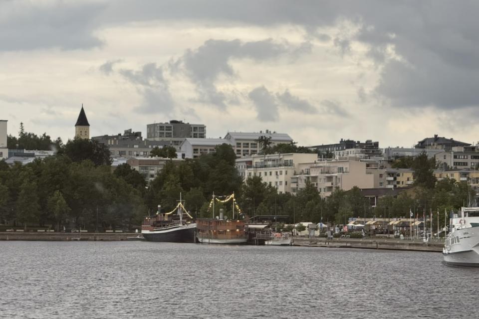 Harborfront in Lappeenranta, Finland, with boats docked along the shoreline and apartment buildings rising behind under a cloudy summer sky.