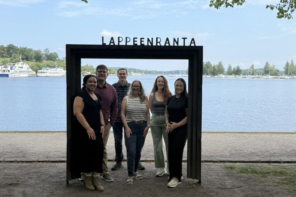 "Six people stand smiling inside a large black rectangular frame with the word 'Lappeenranta' on top. Behind them is a scenic lakeside view with boats, trees, and a partly cloudy sky