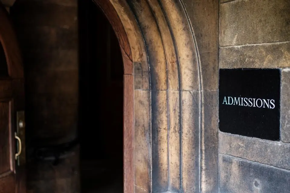 An arched stone doorway with a partially open wooden door leading into a dimly lit space. A black sign on the wall reads “Admissions” in white letters.