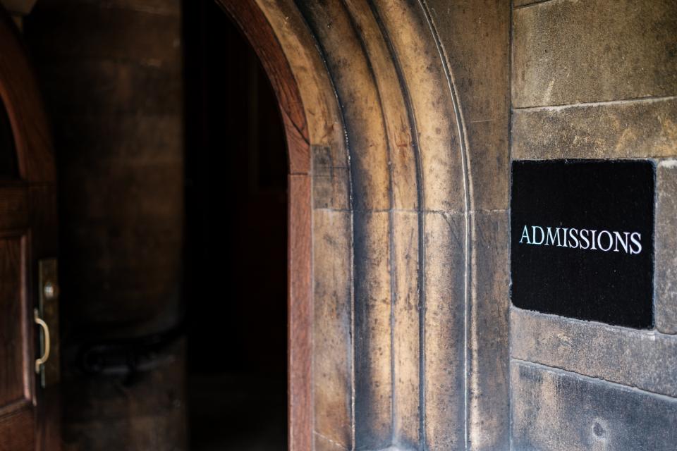 An arched stone doorway with a partially open wooden door leading into a dimly lit space. A black sign on the wall reads “Admissions” in white letters.