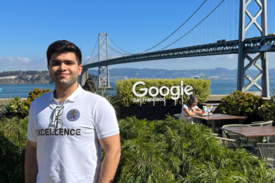 Talha Amin stands on an outdoor terrace at Google’s San Francisco office, with the Bay Bridge and blue sky in the background. He’s wearing a white polo shirt, light pants and sunglasses, surrounded by lush green plants.