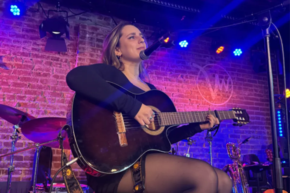 Katya Fomin sitting on a stool on a stage, playing a guitar.