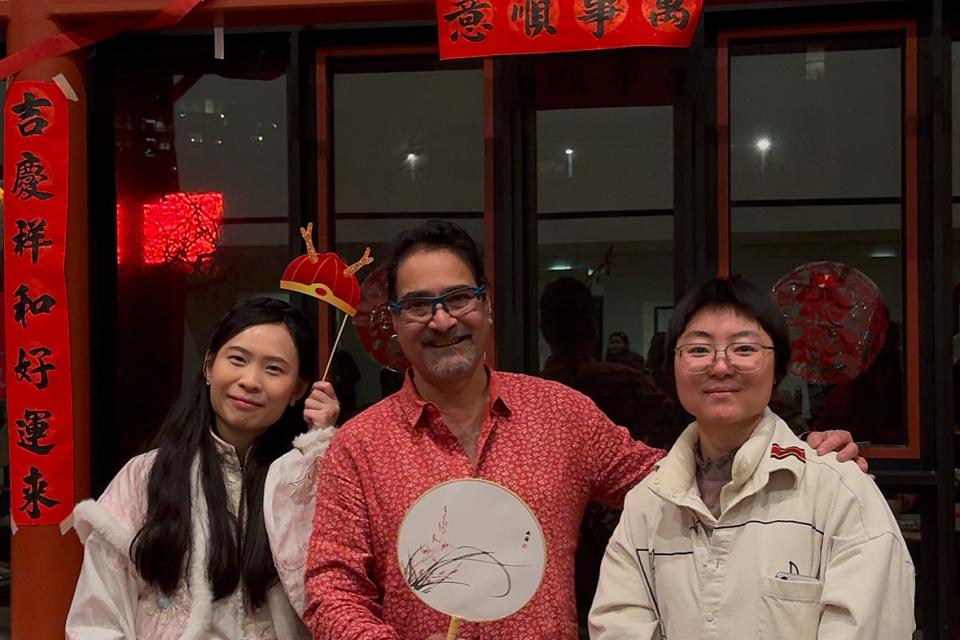 Three people pose indoors at a festive event; two wear traditional clothing, one in modern attire holding a fan. Red decorations and calligraphy frame the window behind them.
