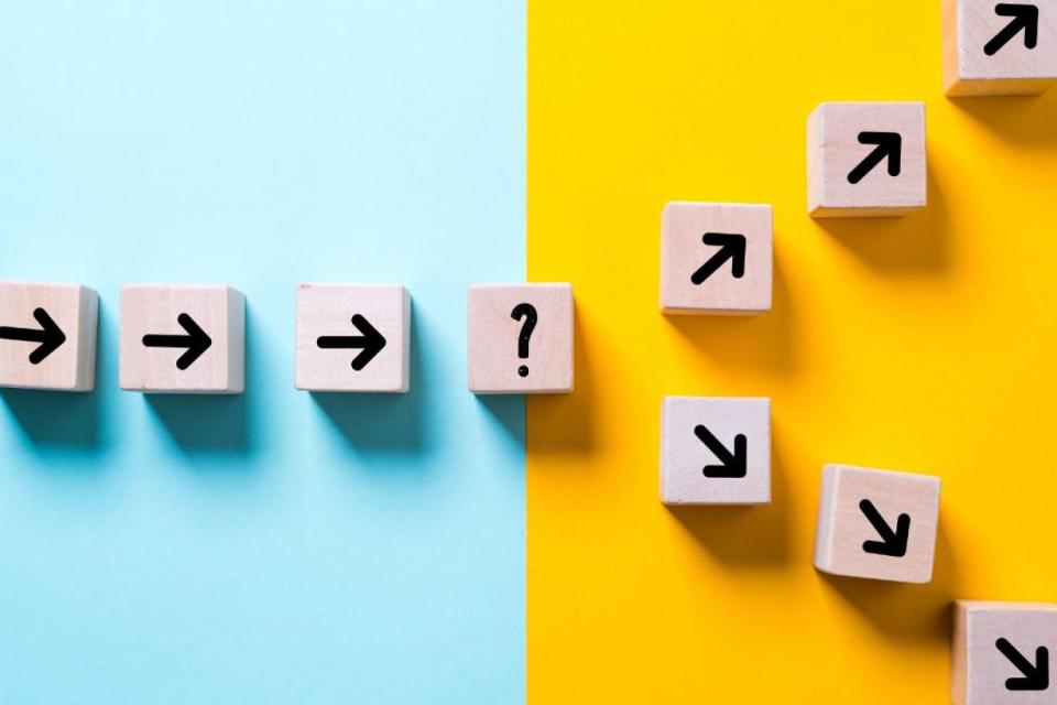 Wooden blocks with black arrows and a question mark form a path, transitioning from a light blue background to a yellow background with arrows going upward.
